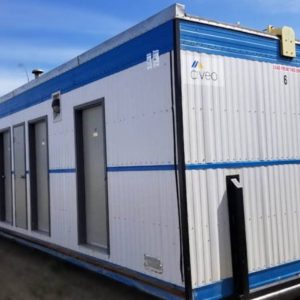 The Lodging Base Camp, a spacious white and blue modular building with multiple doors and windows, features the Civeo logo and is set up outdoors on a dirt surface under a blue sky—perfect for base camp lodging.