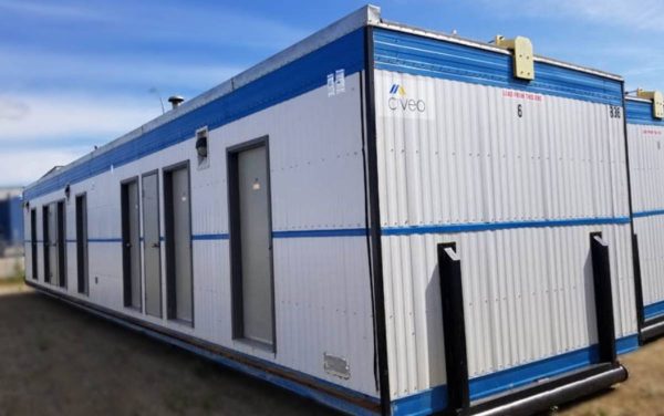 The Lodging Base Camp, a spacious white and blue modular building with multiple doors and windows, features the Civeo logo and is set up outdoors on a dirt surface under a blue sky—perfect for base camp lodging.