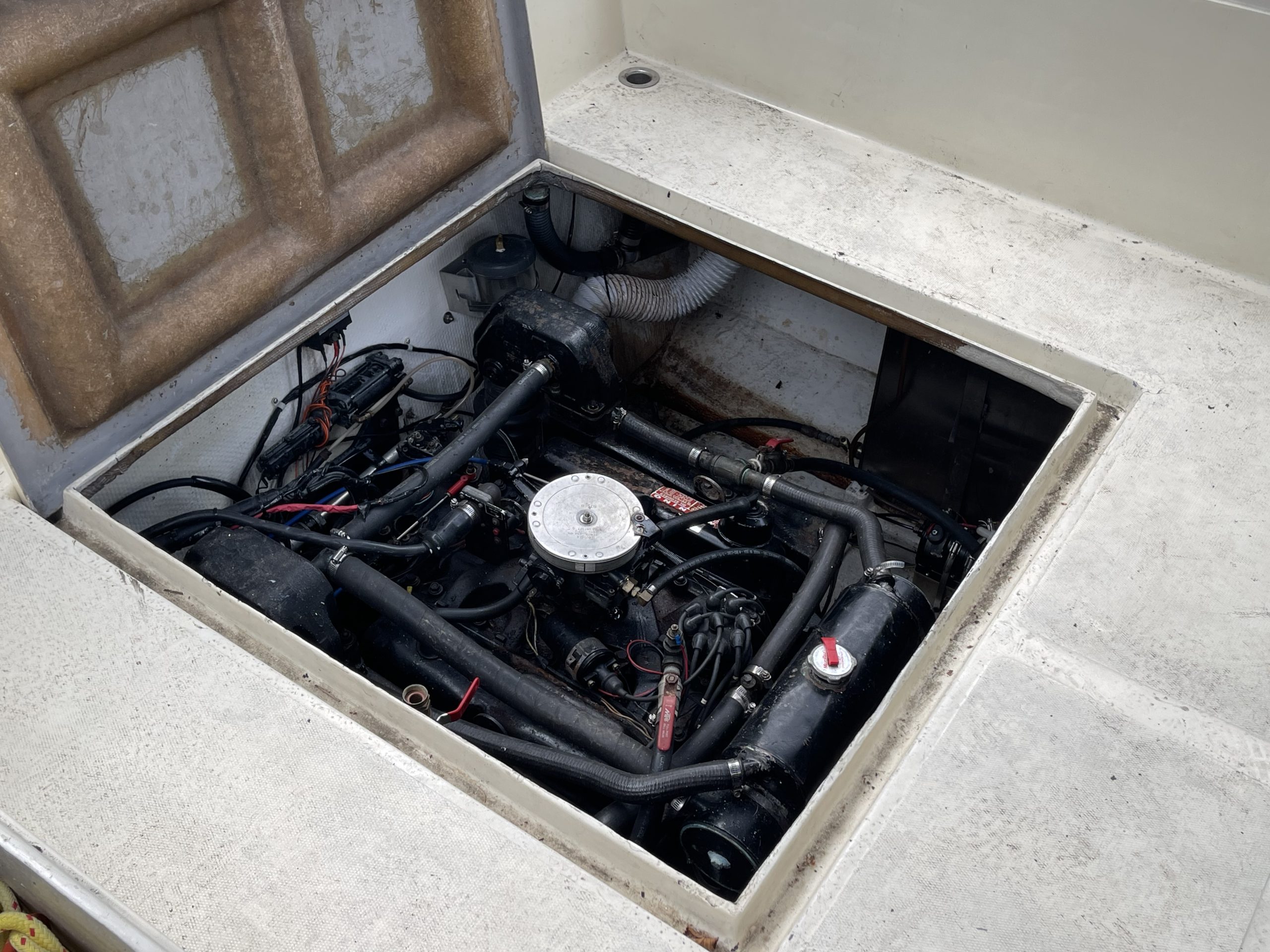 With the hatch open, the engine compartment of the 1976 / 26FT Telestar Sedan is revealed, showcasing a centrally located engine surrounded by an array of black hoses, wires, and metal components neatly set within the classic boat’s white fiberglass deck. The engine appears well-maintained in this vintage vessel.