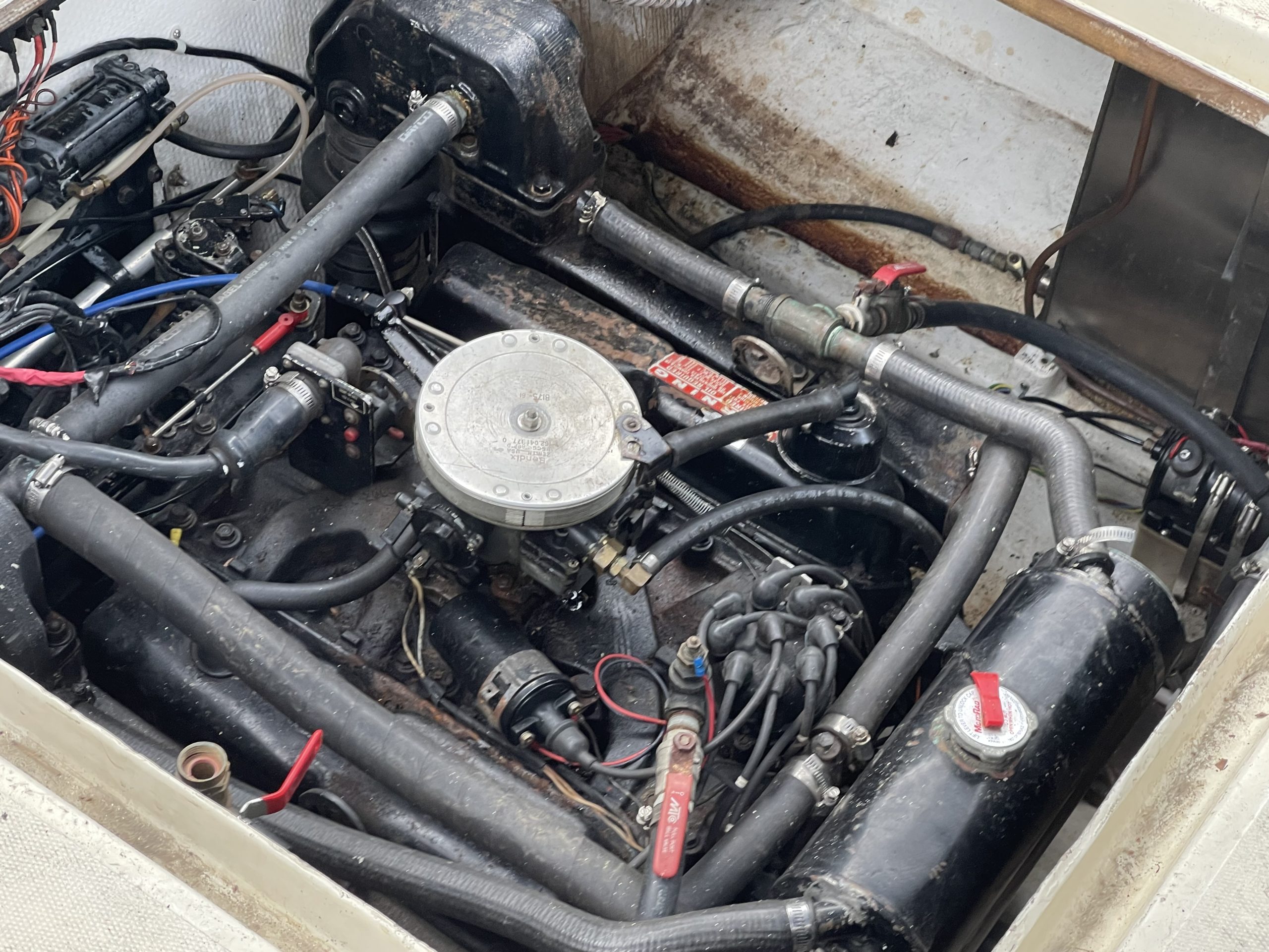 Close-up view of the engine compartment of a 1976 / 26FT Telestar Sedan, showing an array of hoses, wires, and various components. The engine features labeled fittings and some hoses are equipped with red valve handles. Surfaces display evidence of use and weather exposure.