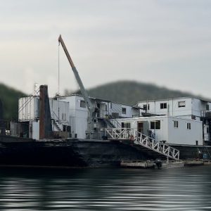 The 21 Person Float Camp features a large barge with white structures and a crane, offering group camping for up to 21 on calm waters, surrounded by forested hills beneath a cloudy sky.