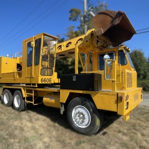A large yellow 660E utility truck with an attached digging bucket is parked on grass near a road, with trees and power lines in the background under a clear blue sky.