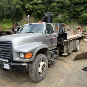 A gray flatbed work truck with a mounted crane is parked on a dirt lot, surrounded by greenery and construction materials. The truck has a visible license plate and company markings on the door.