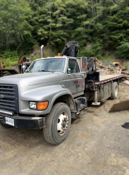 A gray flatbed work truck with a mounted crane is parked on a dirt lot, surrounded by greenery and construction materials. The truck has a visible license plate and company markings on the door.