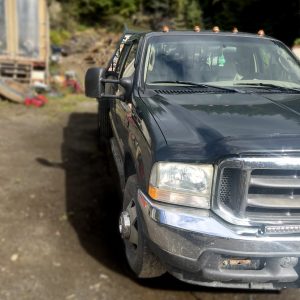 A 2003 Ford F350 Flat Bed Truck, slightly dirty, is parked on a dirt lot with trees and scattered equipment in the background. The image highlights the front and driver's side of this dark-colored flat bed truck.