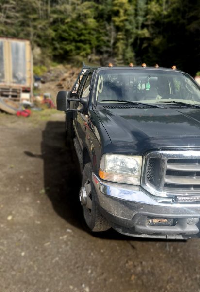A 2003 Ford F350 Flat Bed Truck, slightly dirty, is parked on a dirt lot with trees and scattered equipment in the background. The image highlights the front and driver's side of this dark-colored flat bed truck.