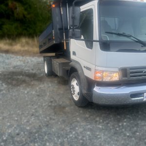 A white dump truck with a raised black bed is parked on a gravel surface, with trees and grass in the background.