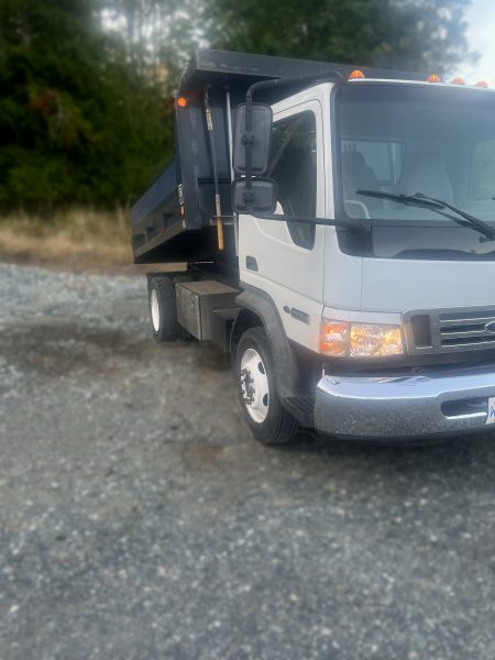 A white dump truck with a raised black bed is parked on a gravel surface, with trees and grass in the background.