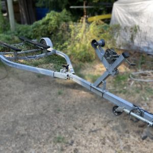 A 2009 Highliner Single Axel Boat Trailer with two wheels, a front winch, and boat support structures is parked on dry ground, with green foliage and outdoor items blurred in the background.
