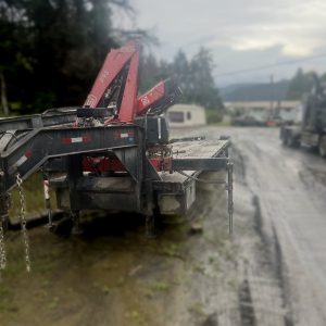 A 2009 Oasis 40ft Flat Deck / HIAB Picker with red hydraulic cranes is parked on a muddy road near trees and buildings under a cloudy sky.