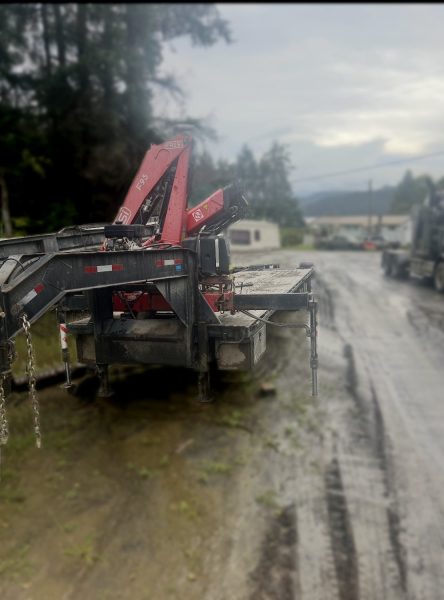 A 2009 Oasis 40ft Flat Deck / HIAB Picker with red hydraulic cranes is parked on a muddy road near trees and buildings under a cloudy sky.