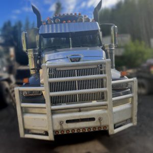 A 2010 Western Star Highway Hauler is parked outdoors on a dirt surface, featuring a silver exterior and front grille guard, with trees and a blue sky with clouds in the background.