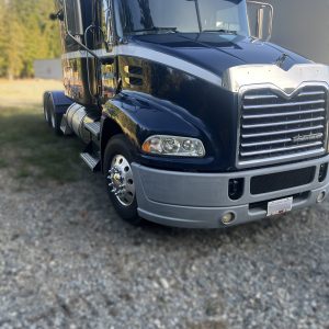 A 2011 Mack Truck with a shiny black finish is parked on gravel by a building, surrounded by trees and grass on a sunny day.