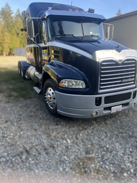 A 2011 Mack Truck with a shiny black finish is parked on gravel by a building, surrounded by trees and grass on a sunny day.