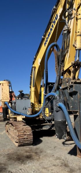 A 2013 PC300 Komatsu Hoe Drill in yellow with blue hoses is parked on a construction site under a clear sky, featuring large metal tracks, hydraulic components, and Komatsu's renowned reliability.