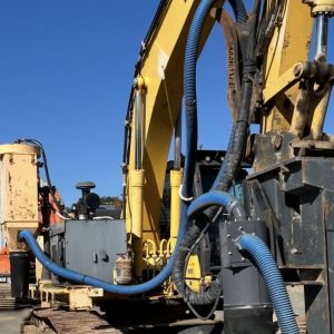 A 2013 PC300 Komatsu Hoe Drill in yellow with blue hoses is parked on a construction site under a clear sky, featuring large metal tracks, hydraulic components, and Komatsu's renowned reliability.