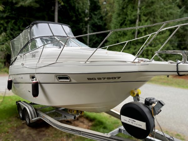A 1997 / 24FT Maxum SCR with a clear canopy is parked on a trailer near a road, trees in the background. Its registration number is visible along the hull.