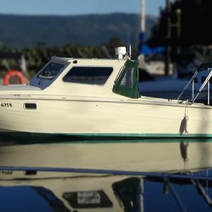 A small cream-colored Sea Ray motorboat with a covered cabin and custom refit is docked on calm water at a marina, reflecting clearly on the surface. Other boats and docks are visible in the blurred background.