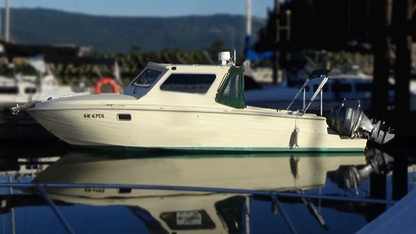 A small cream-colored Sea Ray motorboat with a covered cabin and custom refit is docked on calm water at a marina, reflecting clearly on the surface. Other boats and docks are visible in the blurred background.