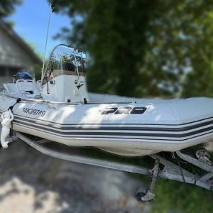 A 2001 / 18FT Zodiac Open Pro RHIB with black stripes and registration 14K39749 is mounted on a trailer, parked outdoors near trees and a building. "PRO" is visible on the side of the white motorboat.