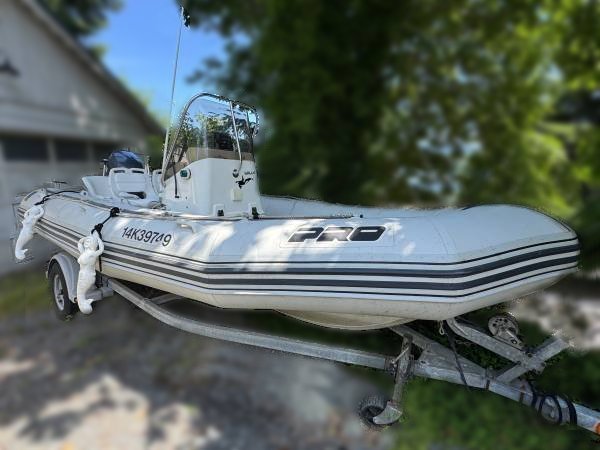 A 2001 / 18FT Zodiac Open Pro RHIB with black stripes and registration 14K39749 is mounted on a trailer, parked outdoors near trees and a building. "PRO" is visible on the side of the white motorboat.