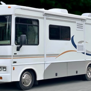 A white Winnebago motorhome with blue and tan stripes is parked on a paved road, surrounded by green trees in the background.