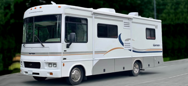 A white Winnebago motorhome with blue and tan stripes is parked on a paved road, surrounded by green trees in the background.