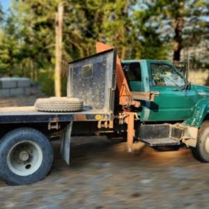 A 1993 GMC Top Kick Flat Bed with HIAB Crane and a spare tire on the back is parked on a dirt road, surrounded by trees and a wooded area.