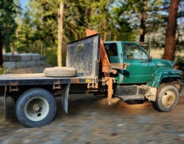 A 1993 GMC Top Kick Flat Bed with HIAB Crane and a spare tire on the back is parked on a dirt road, surrounded by trees and a wooded area.