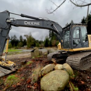A yellow and black 2013 John Deere 225D IC excavator is parked outdoors on a cloudy day, its large metal bucket resting near several big rocks with trees in the background.