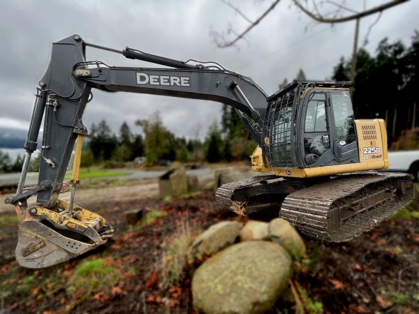 A yellow and black 2013 John Deere 225D IC excavator is parked outdoors on a cloudy day, its large metal bucket resting near several big rocks with trees in the background.