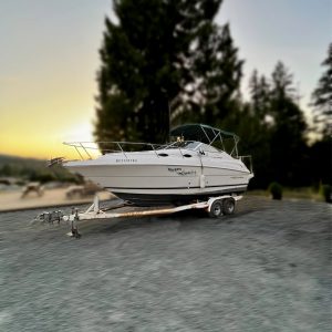A 2000 / 24FT Monterey 242 boat sits on a trailer parked on gravel, surrounded by trees and a sunset in the background.