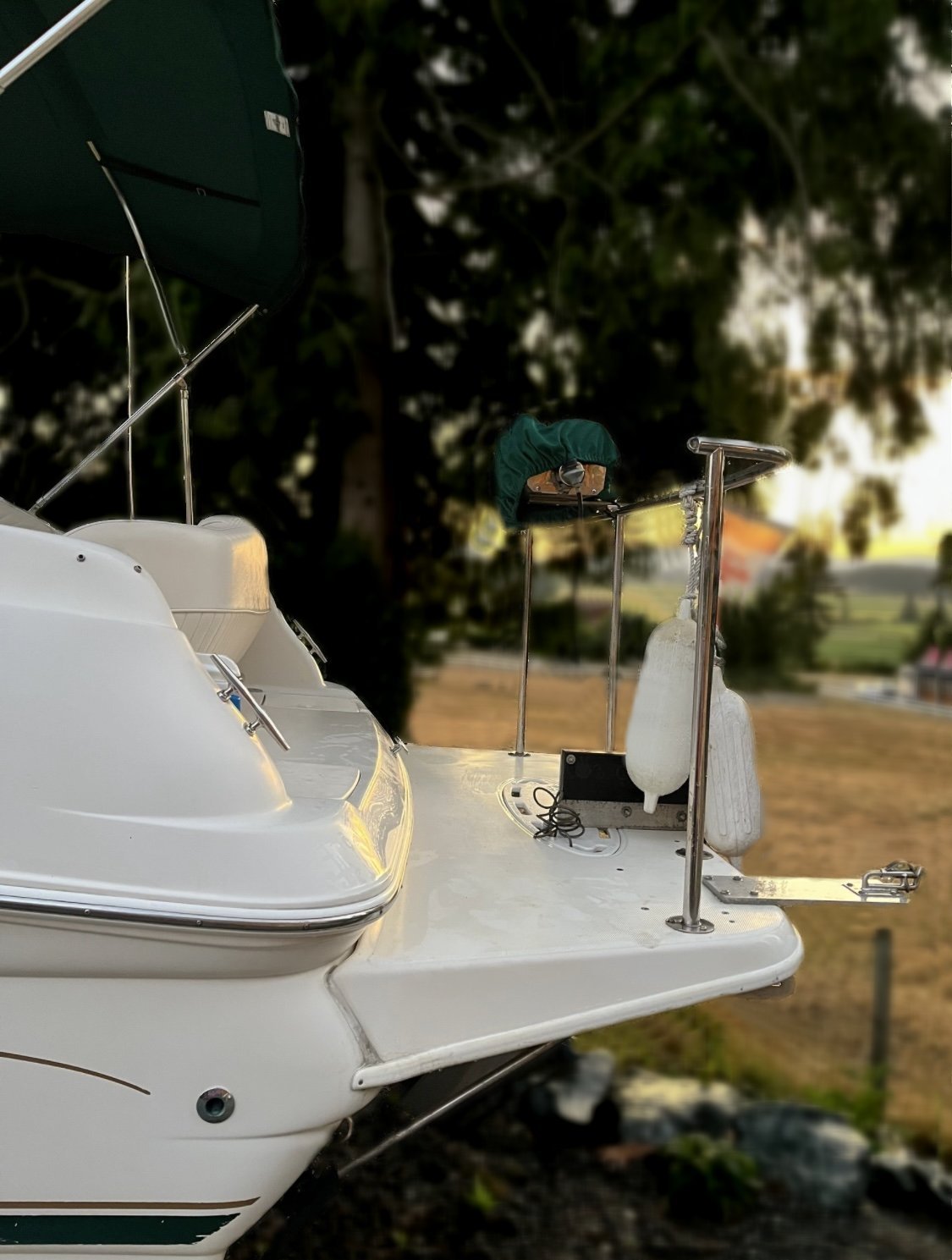 A close-up of the rear deck of a white 2000 / 24FT Monterey 242 boat with a green canopy, metal railings, rope fender, and equipment, docked or on land, with trees and grass visible in the background.