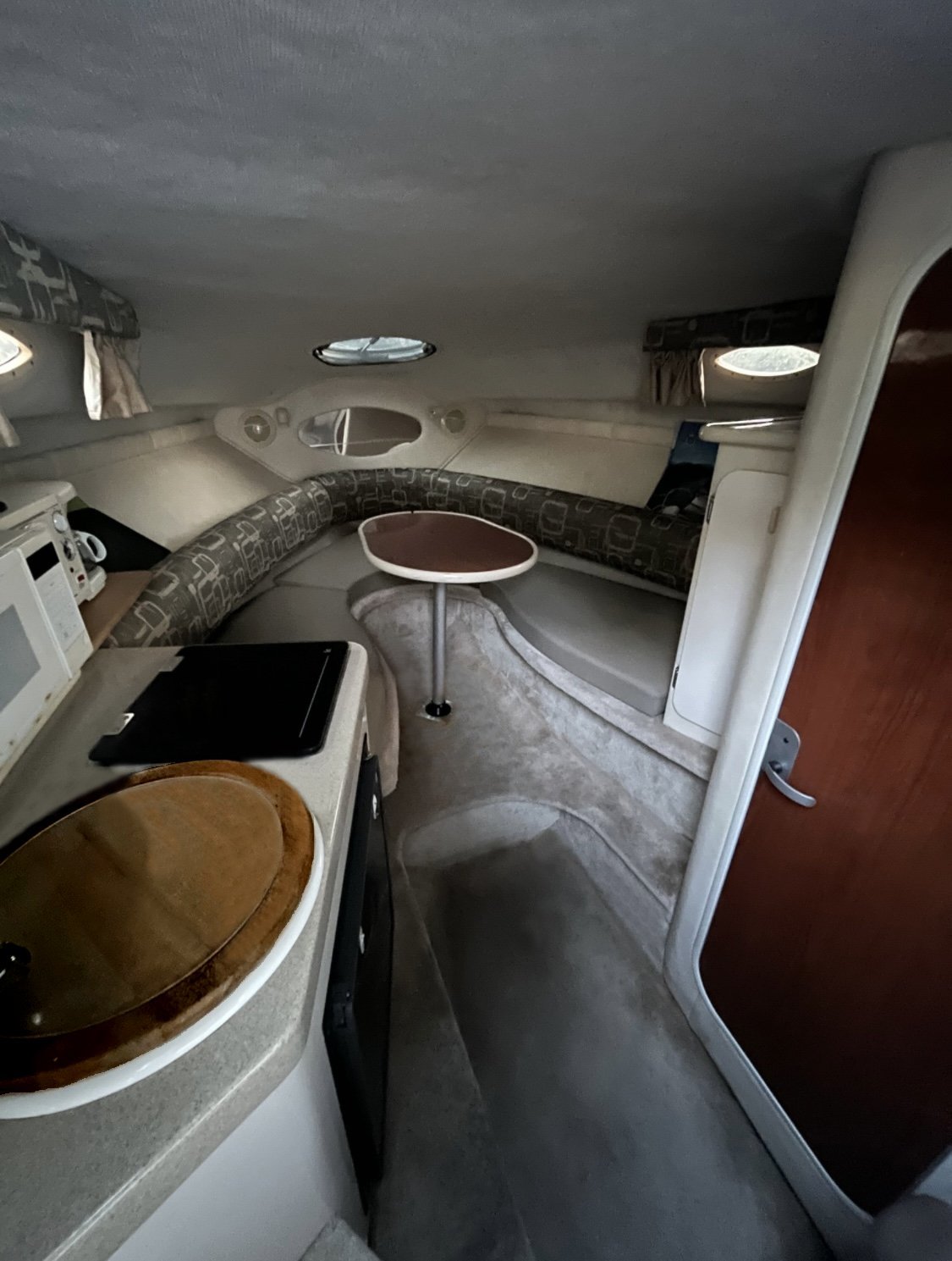 Interior of a 2000 / 24FT Monterey 242 cabin featuring curved seating with patterned cushions, a round table, compact kitchenette with sink, laptop on the counter, and a closed wooden door to the right in this cozy space.