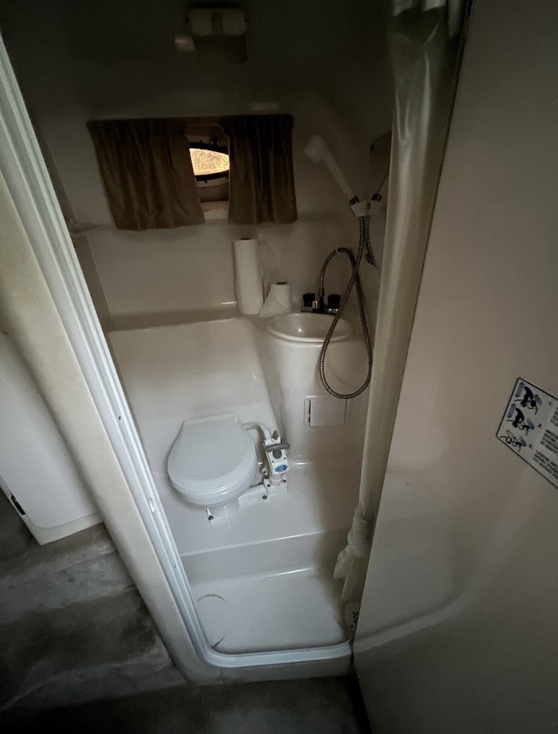 The 2000 / 24FT Monterey 242 RV bathroom includes a white toilet, small sink, handheld showerhead, and curtained window above. Light walls and a partially open door enclose this compact space.
