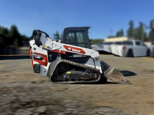 A 2023 Bobcat T66 Skidsteer in white and orange with a front bucket is parked on dirt, with blurred vehicles and trees visible in the background.