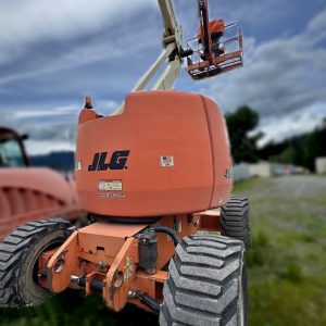 A 2008 JLG 450AJ Man Lift with thick tires is parked on grass outdoors. Its orange cage, holding a person, is raised high against a cloudy sky, with construction equipment visible in the background.