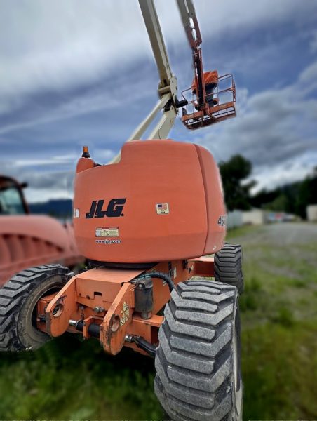 A 2008 JLG 450AJ Man Lift with thick tires is parked on grass outdoors. Its orange cage, holding a person, is raised high against a cloudy sky, with construction equipment visible in the background.