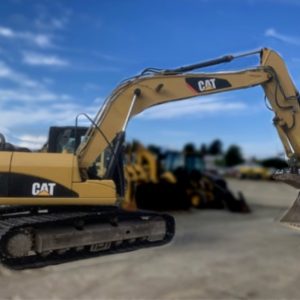 A yellow 2011 CAT 3150 Excavator with a large bucket is parked on a dirt lot, while other construction vehicles appear blurred in the background under a clear blue sky.