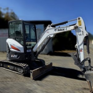 A 2021 Bobcat E26 Excavator Mini Hoe in white and black with a digging bucket is parked on gravel, with other construction equipment and trees blurred in the background.