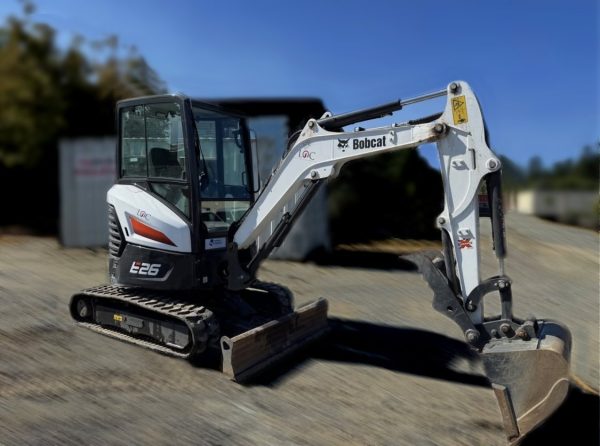 A 2021 Bobcat E26 Excavator Mini Hoe in white and black with a digging bucket is parked on gravel, with other construction equipment and trees blurred in the background.