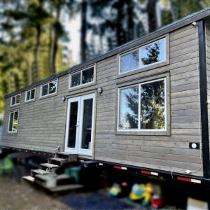 The 2021 is a modern tiny house on wheels featuring wood siding, large windows, and a glass double door, stationed outdoors in the woods with stairs up to the entrance.