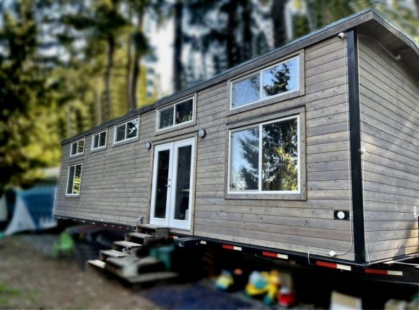 The 2021 is a modern tiny house on wheels featuring wood siding, large windows, and a glass double door, stationed outdoors in the woods with stairs up to the entrance.