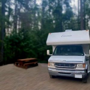 A 1999 28FT Ford Corsair camper van is parked at a forest campsite beside a wooden picnic table, surrounded by tall trees and lush greenery.