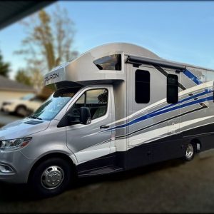 A 2024 / 24FT Thor Tiburon Motor Coach in silver and blue is parked on a suburban street, featuring a large windshield and spacious side awning, with houses and trees in the background.
