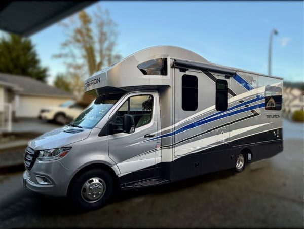 A 2024 / 24FT Thor Tiburon Motor Coach in silver and blue is parked on a suburban street, featuring a large windshield and spacious side awning, with houses and trees in the background.