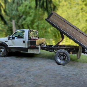 A white 2000 Ford F450 Dump Truck with its wooden flatbed lifted as if unloading is parked on a gravel road, with green trees in the background.