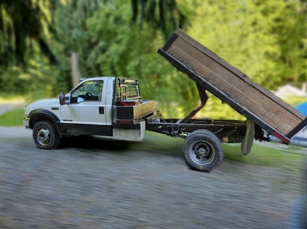 A white 2000 Ford F450 Dump Truck with its wooden flatbed lifted as if unloading is parked on a gravel road, with green trees in the background.