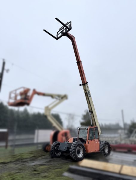 A red and yellow telescopic forklift with an extended boom arm and fork attachment is parked outdoors on grass, with another boom lift visible in the blurred background on a cloudy day.