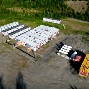 Aerial view of a remote site with 1998 / Relocatable Industrial Accommodation—rectangular buildings, trailers, propane tanks, and trucks on a gravel lot—surrounded by dense green forest.